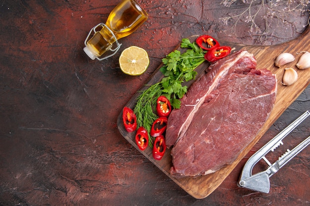 Oben Blick auf rotes Fleisch auf Holzbrett und Knoblauch grün gehackter Pfeffer gefallene Ölflasche Zitrone auf dunklem Hintergrund