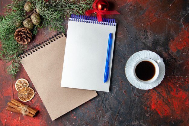 Oben blick auf spiralnotizbücher mit stift-tannenzweig-zimt-limonen-koniferenkegel und einer tasse schwarzem tee auf dunklem hintergrund