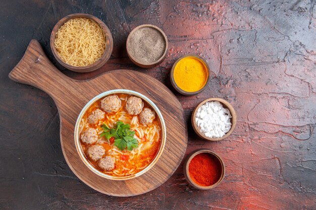 Oben Blick auf Tomaten-Fleischbällchen-Suppe mit Nudeln in einer braunen Schüssel und verschiedenen Gewürzen auf dunklem Hintergrund