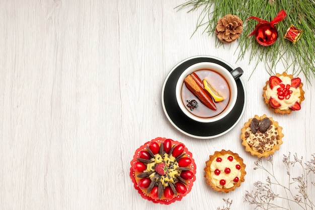 Oben entfernte Ansicht eine Tasse Zitronen-Zimt-Tee-Beeren-Kuchen-Törtchen und die Kieferblätter mit Weihnachtsspielzeug auf der rechten Seite des weißen Holzbodens