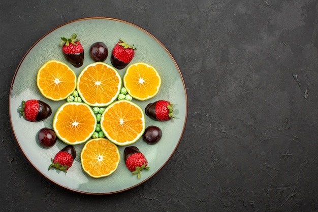 Kostenloses Foto oben nahaufnahme früchte und schokolade gehackte orange mit schokoladenüberzogenen erdbeeren und grünen bonbons auf der linken seite des dunklen tisches