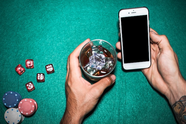 Obenliegende Ansicht der Hand des Mannes, die Mobiltelefon- und Whiskyglas über der Pokertabelle hält