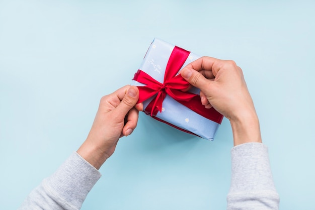 Obenliegende Ansicht der Hand roten Bandbogen auf Geschenkbox über blauem Hintergrund binden