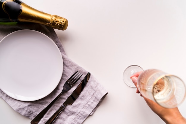 Obenliegende ansicht der menschlichen hand champagnerglas nahe leerer weißer platte auf einfachen hintergrund setzend