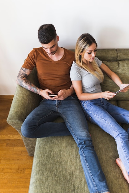 Obenliegende Ansicht von den Paaren, die auf Couch unter Verwendung der elektronischen Geräte sitzen