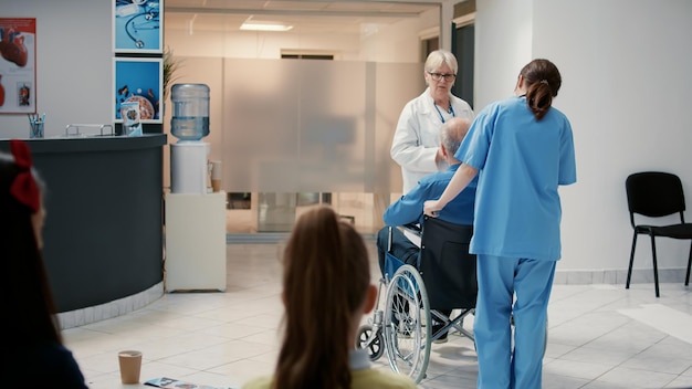 Oberarzt spricht mit pensioniertem Mann mit Behinderung an der Krankenhausrezeption und bereitet sich auf ärztliche Beratung vor. Rollstuhlfahrer in der Lobby des Wartezimmers, der an einem Kontrolltermin teilnimmt.