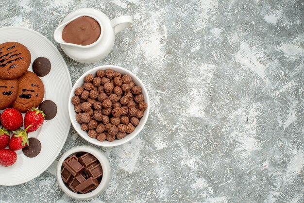 Obere Hälfte Ansicht Schokoladenkekse Erdbeeren und runde Pralinen auf dem weißen ovalen Teller und Schalen mit Pralinen Müsli und Kakao auf dem grau-weißen Grund