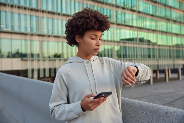 Oberkörperaufnahme einer aktiven lockigen jungen Frau nutzt Telefonanwendung und Smartwatch überprüft Fitnessergebnisse trägt lässige Hoodie-Posen gegen verschwommenes Glas urbanes Gebäude Lifestyle-Konzept