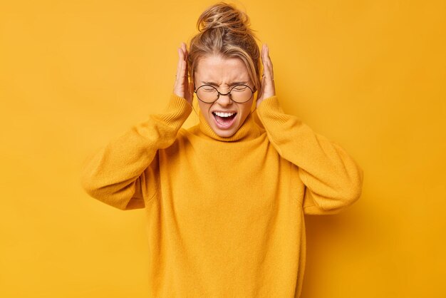 Oberkörperaufnahme einer emotionalen jungen blonden Frau, die laut schreit, die Ohren mit den Händen bedeckt, will kein störendes Geräusch hören, trägt eine Brille und einen Pullover, die über gelbem Hintergrund isoliert sind. Stoppen Sie dies
