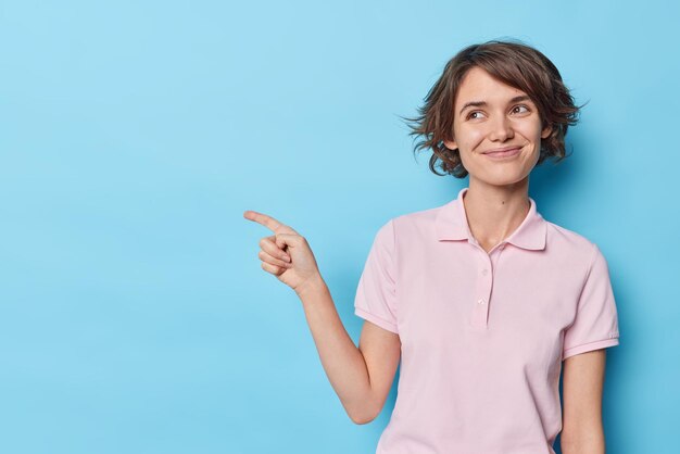 Oberkörperaufnahme einer erfreuten jungen hübschen Frau mit Bob-Frisurlächeln, die glücklich ein lässiges T-Shirt trägt, das auf einen leeren Kopierraum für Ihre Werbung zeigt, die über blauem Hintergrund isoliert ist. Schau dir das an