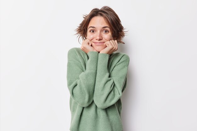 Oberkörperaufnahme einer schönen Frau, die die Hände unter dem Kinn hält, lächelt glücklich und sieht neugierig auf etwas aus, das einen lässigen, warmen Pullover trägt, der über weißem Hintergrund isoliert ist. Erfreutes fröhliches weibliches Modell drinnen