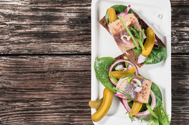 Offene Sandwiches mit jungem Hering, Kräutern und eingelegter Gurke auf einem weißen Teller, Draufsicht mit Kopienraum. Traditionelles dänisches Smorrebrod. Sandwiches mit Hering sind auf einem alten Holztisch angeordnet