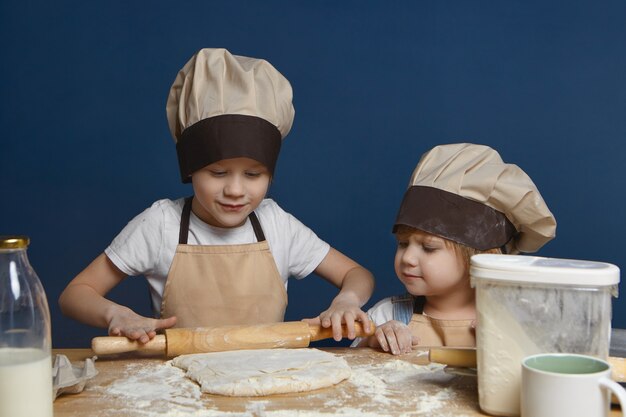 Offener Schuss des charmanten kleinen Mädchens in der Kochmütze, die ihren älteren Bruder beobachtet, der Teig für Kekse oder Kuchen knetet