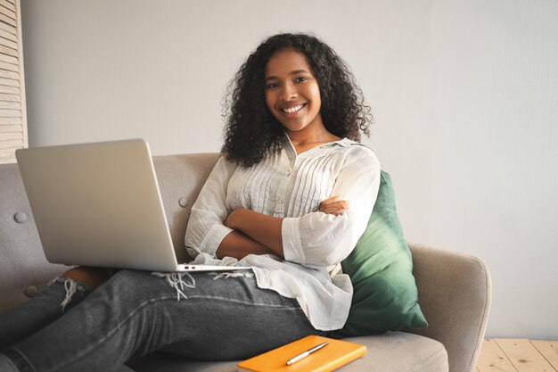 Offener Schuss des glücklichen erfolgreichen jungen dunkelhäutigen weiblichen Bloggers, der auf Sofa mit modernem elektronischem Gerät auf ihrem Schoß sitzt, Arme verschränkt hält und selbstbewusst lächelt, Internet surft