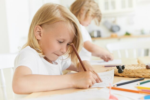 Offenes Porträt von zwei Vorschulkindern, die Freizeit drinnen zu Hause oder im Kindergarten verbringen, die zusammen am hölzernen Schreibtisch mit Bleistiften und Blatt Papier sitzen, Zeichnung. Entwicklung und Kreativität