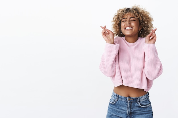 Oh Gott, bitte lass es geschehen. Porträt eines hoffnungsvoll aufgeregten und glücklichen, charmanten afroamerikanischen B-Day-Mädchens, das die Augen wünscht und freudig lächelt, als Kreuzfinger für viel Glück über der weißen Wand