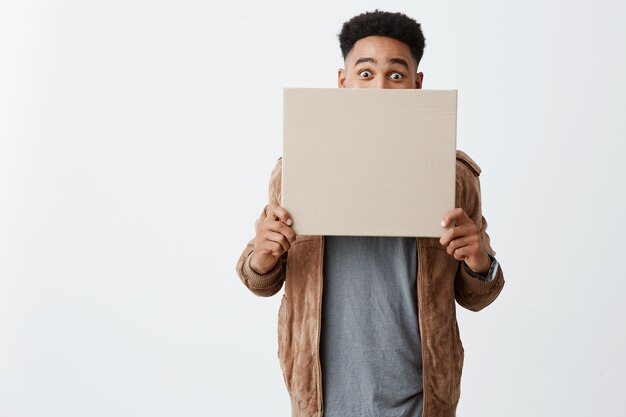 OH MEIN GOTT. Nahaufnahme des jungen schönen lustigen schwarzhäutigen Mannes mit Afro-Frisur im Gre-Shirt unter braunem Jackenschließgesicht mit Karton, Blick in die Kamera mit überraschtem Ausdruck