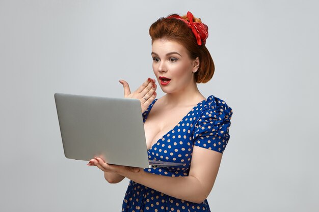 Oh mein Gott. Studioaufnahme der herrlichen emotionalen jungen Dame im Weinlese-Outfit, das generischen Laptop-Computer hält, überraschten Ausdruck, Handfläche an ihrem Mund haltend
