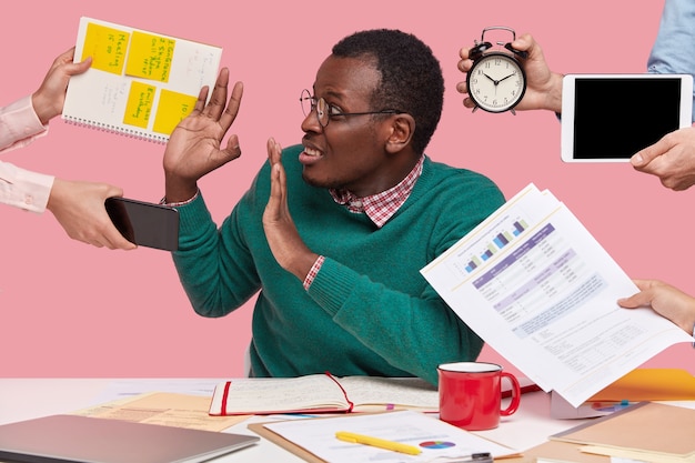 Oh nein, nicht jetzt! Gestresster dunkelhäutiger Mann hat viel Arbeit im Büro, gefragt von vielen Leuten auf einmal, die an die Vorbereitung eines Geschäftsprojekts erinnern, Wecker halten