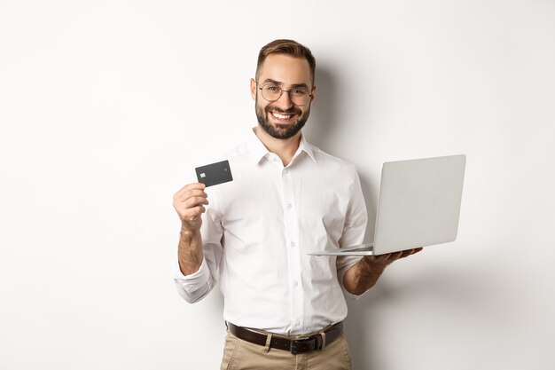 Online Einkaufen. Hübscher Mann, der Kreditkarte zeigt und Laptop verwendet, um im Internet zu bestellen, stehend