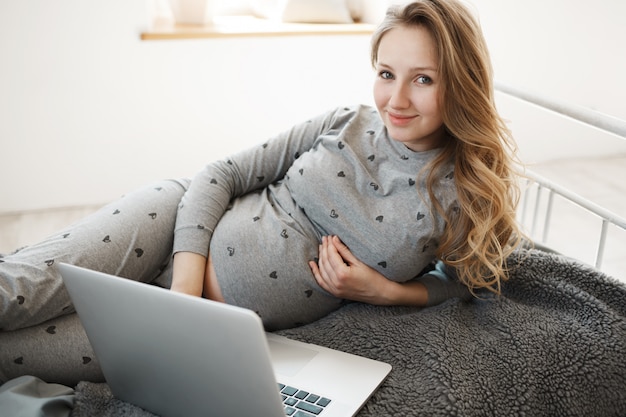 Online einkaufen. porträt der glücklichen jungen schwangeren frau mit hellem haar in der hauskleidung, die auf bett liegt und versucht, kleidung für zukünftiges baby im online-shop auf laptop-computer zu finden.