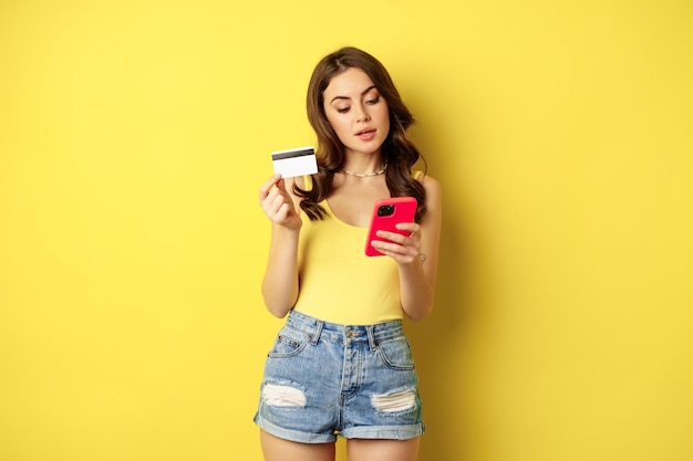 Online Einkaufen. Stilvolle brünette Frau, die Smartphone und Kreditkarte hält, in App bezahlt, Handyanwendung verwendet, etw kauft, über gelbem Hintergrund steht