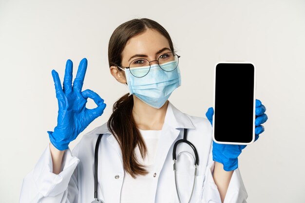 Online-Konzept der medizinischen Hilfe Begeisterte junge Ärztin mit Gesichtsmaske, die ein Okay-Zeichen und ein Handy zeigt ...