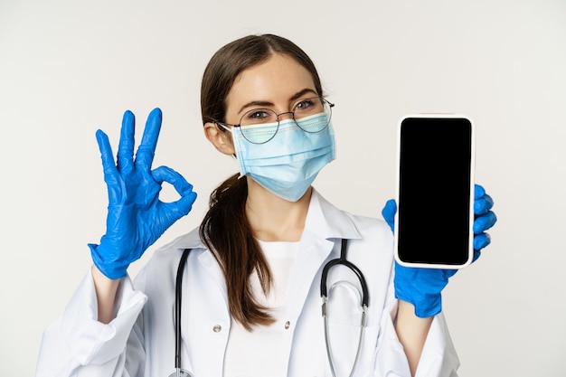 Online-Konzept der medizinischen Hilfe Begeisterte junge Ärztin mit Gesichtsmaske, die ein Okay-Zeichen und ein Handy zeigt ...