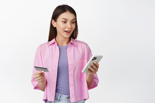 Online-Shopping Aufgeregte junge Frau, die eine Kreditkarte hält und überrascht über die Bestellung einer Handy-App auf Antrag steht, die über weißem Hintergrund steht