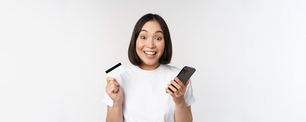 Online-Shopping Fröhliche asiatische Frau mit Kreditkarte und Smartphone-App, die auf der Website über den weißen Hintergrund des Mobiltelefons bezahlt