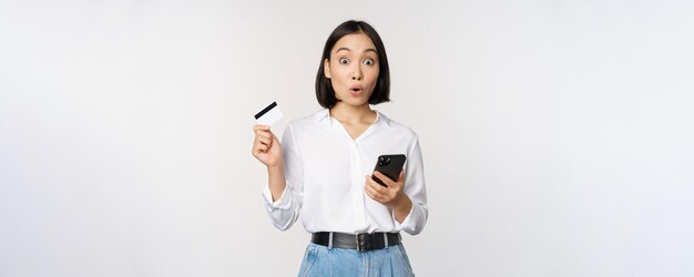 Online-Shopping-Konzept Bild eines überraschten asiatischen Mädchens mit Kreditkarte und Smartphone, das ungläubig auf den weißen Hintergrund der Kamera schaut