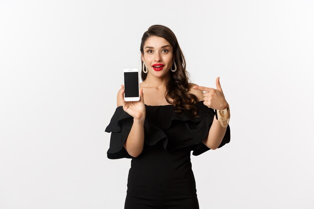 Online-Shopping-Konzept. Modische Frau im schwarzen Kleid, das Finger auf Smartphonebildschirm zeigt, Anwendung zeigt, über weißem Hintergrund stehend.