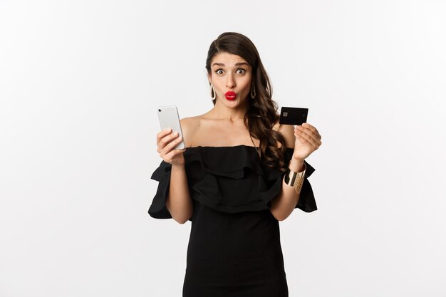 Online-Shopping-Konzept. Modische Frau im schwarzen Kleid, die Kreditkarte mit Smartphone hält, aufgeregt aussieht und auf weißem Hintergrund steht