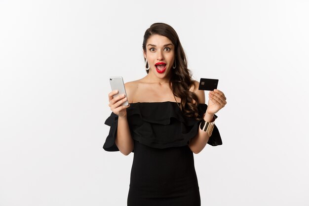 Online-Shopping-Konzept. Modische Frau im schwarzen Kleid, Kreditkarte mit Smartphone haltend, aufgeregt schauend, über weißem Hintergrund stehend.