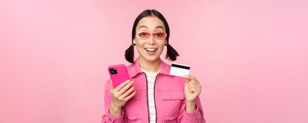 Online-Shopping Lächelnde asiatische Käuferin mit Smartphone und Kreditkarte, die in der mobilen App bezahlt, die über rosafarbenem Hintergrund steht