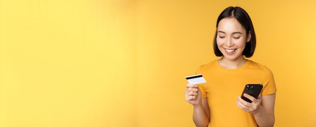 Online-Shopping Lächelndes asiatisches Mädchen, das Kreditkarte und Handy-App verwendet, um kontaktlose Bestellung auf Smartphone-Anwendung zu bezahlen, die über gelbem Hintergrund steht