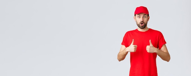 Online-Shopping-Lieferung während Quarantäne und Takeaway-Konzept Beeindruckter und schockierter bärtiger Kurier in rotem T-Shirt und Mützenuniform, der mit offenem Mund nach Luft schnappt, erstaunt Daumen hoch, um den Service zu empfehlen