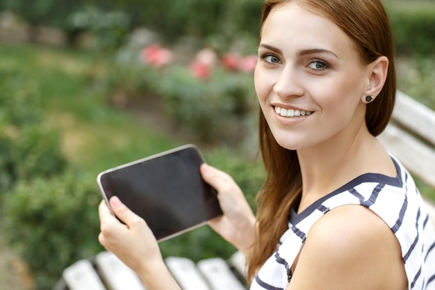 Online-Shopping Schöne Frau, die mit einem Lächeln auf die Kamera über die Schulter schaut und einen im Park sitzenden Tisch hält