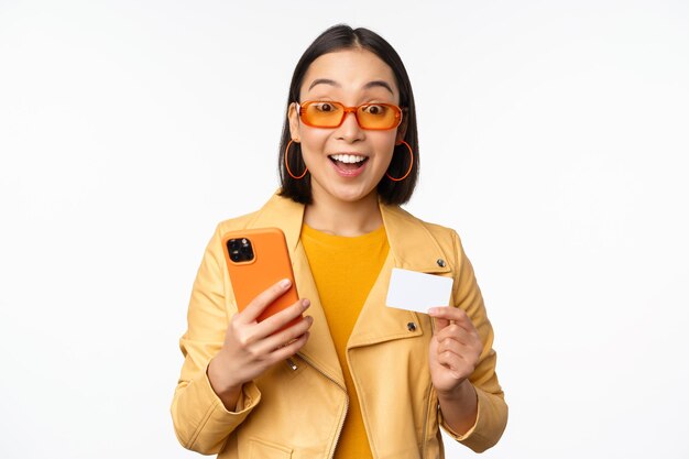 Online-Shopping Stilvolles asiatisches weibliches Modell mit Sonnenbrille, das Kreditkarte und Handy hält und glücklich stehend über weißem Hintergrund lächelt Kopieren Sie Platz