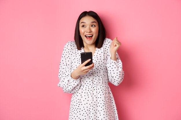 Online-Shopping und Beauty-Konzept. Aufgeregte asiatische Frau, die im Internet gewinnt, Smartphone hält und sich freut, glücklich lächelt und feiert, auf rosa Hintergrund steht
