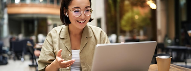 Kostenloses Foto online-treffen einer jungen asiatischen frau aus der stadt, die mit einem laptop-video-chat spricht und gestikuliert, während sie in einem sitzt