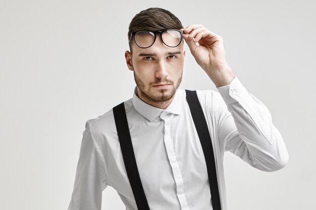 Optik, Brille und Modekonzept. Bild des hübschen unrasierten jungen brünetten Mannes, der weißes formelles Hemd mit Hosenträgern trägt, mit prüfendem Blick in die Kamera starrt und seine Brille hebt