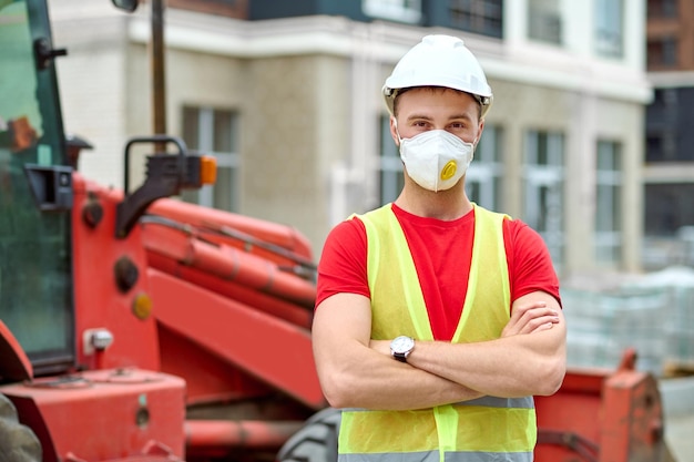Optimismus. Mann in Schutzmaske und Helm in Arbeitsweste mit gefalteten Händen, die gut gelaunt auf der Baustelle am Nachmittag in die Kamera schauen
