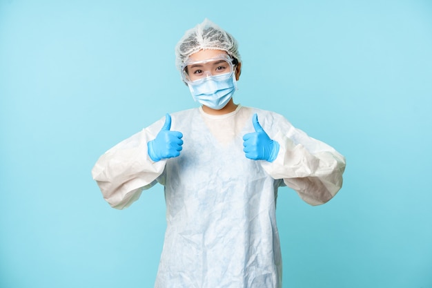 Optimistische asiatische Krankenschwester oder Ärztin, die persönliche Schutzausrüstung trägt, Daumen nach oben zeigt, Kampagne zur Coronavirus-Prävention, blauer Hintergrund.