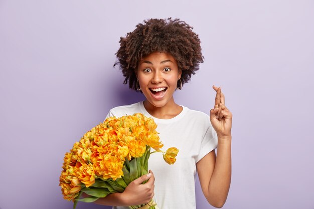 Optimistische Frau glaubt aufrichtig an gutes Wohlbefinden, hebt die Hand mit gekreuzten Fingern, hält schöne gelbe Frühlingsblumen, hat glücklichen Ausdruck, trägt weißes T-Shirt isoliert über lila Wand