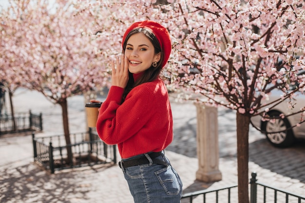 Optimistische Frau im hellen Outfit lächelt niedlich nahe Sakura. Hübsche Dame im roten Pullover und im Hut, der im Stadtgarten in guter Laune aufwirft