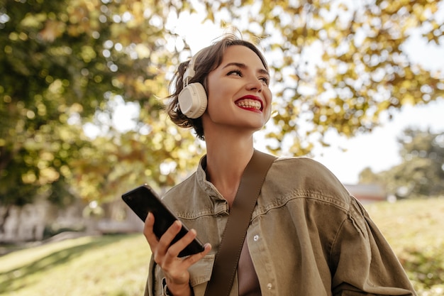 Optimistische Frau mit brünetten Haaren in Denim-Oliven-Kleidern lächelt und hält Telefon draußen. Frau in leichten Kopfhörern posiert im Freien.