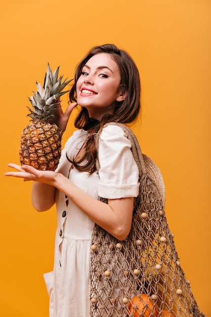 Optimistische Frau mit großer Öko-Tasche, die Ananas hält und auf orange Hintergrund lächelt.