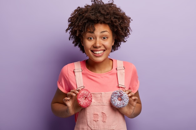 Optimistische fröhliche dunkelhäutige Frau mit Afro-Frisur, hält zwei süße funkelnde Donuts, hat Spaß mit Süßigkeiten