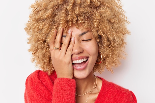 Optimistische, glückliche, lockige Frau bedeckt das Gesicht mit der Hand und lacht freudig über etwas, das in einem lässigen roten Pullover gekleidet ist, fühlt sich überglücklich und drückt positive Emotionen einzeln auf weißem Hintergrund aus.